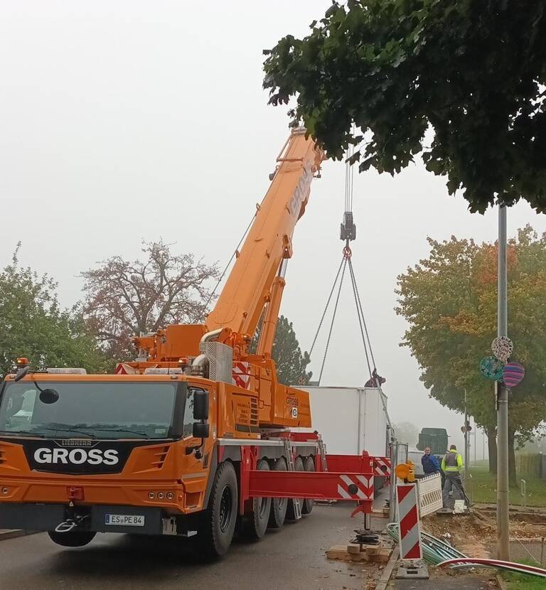 Ein großer orangefarbener Kran hebt ein Objekt auf einer Straße, während Bauarbeiter daneben stehen.