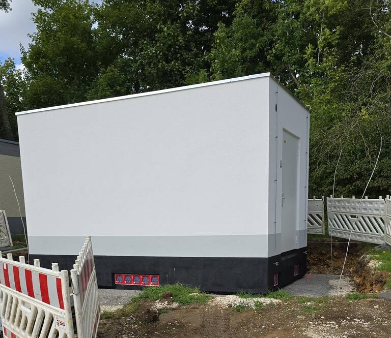 Ein kleines, weißes Gebäude steht in einer Baustelle, umgeben von Bäumen und Absperrungen.