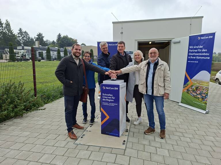 Eine Gruppe von sechs Personen steht lächelnd um eine Säule und berührt gemeinsam einen Knopf, um den regionalen Glasfaserausbau durch NetCom BW zu symbolisieren, während im Hintergrund Roll-Up-Banner zu sehen sind.