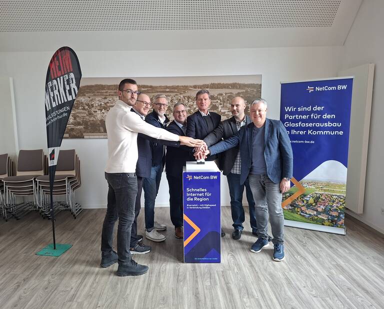 Eine Gruppe von sieben Personen steht lächelnd um eine Säule und berührt gemeinsam einen Knopf, um den regionalen Glasfaserausbau durch NetCom BW zu symbolisieren, während im Hintergrund Roll-Up-Banner und ein Werbefahne zu sehen sind.
