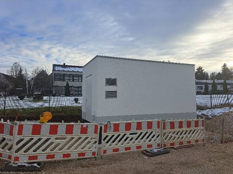 Ein weißes, kubisches Gebäude ist von einer rot-weißen Absperrung umgeben, im Hintergrund sind weitere Häuser und bewölkter Himmel zu sehen.