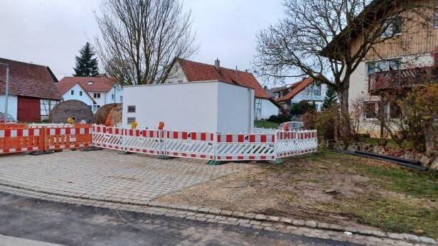 Ein weißes, kubisches Gebäude ist von einer rot-weißen Absperrung umgeben, im Hintergrund sind weitere Häuser und bewölkter Himmel zu sehen.
