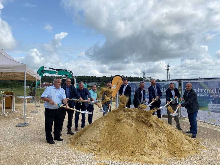 Eine Gruppe von Männern in Anzügen hebt mit Schaufeln Sand auf einer Baustelle, während im Hintergrund ein Bagger zu sehen ist. Spatenstich zum Breitbandausbau
