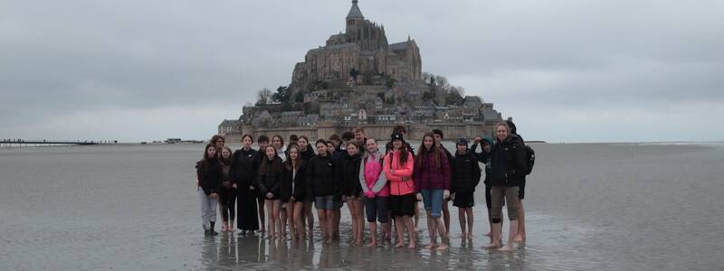 Eine Gruppe von Menschen steht barfuß vor dem berühmten Klosterberg Mont Saint-Michel auf einer nassen Sandfläche.