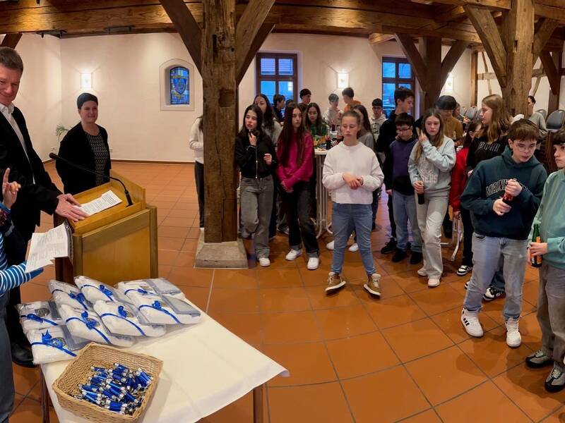 Eine Gruppe von Menschen steht in einem Raum mit Holzsäulen und Ziegelfußboden; vorne links spricht ein Mann hinter einem Pult, daneben eine Frau mit einer Kiste voller Geschenke.