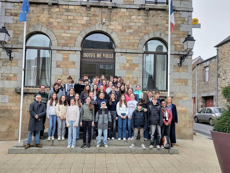 Eine große Gruppe von Menschen posiert lächelnd für ein Foto vor einem Gebäude mit der Aufschrift "Hotel de Ville", flankiert von EU- und französischen Flaggen.