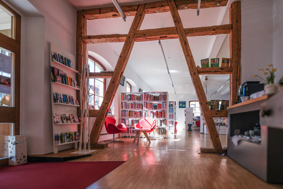 Ein heller, moderner Raum mit Holzbalken, zwei roten Stühlen und Bücherregalen auf einem Holzboden.