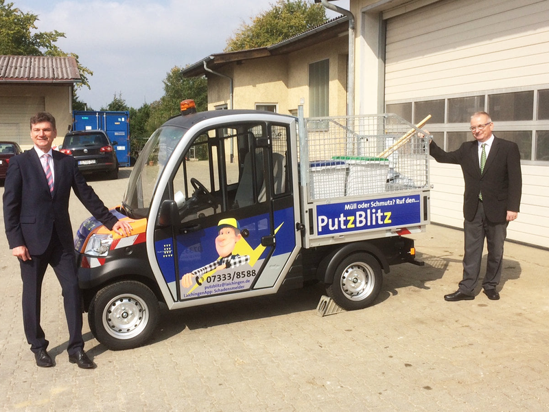 Zwei Männer in Anzügen stehen neben einem kleinen Nutzfahrzeug mit der Aufschrift "PutzBlitz" und einem darauf abgebildeten Cartoon-Charakter.