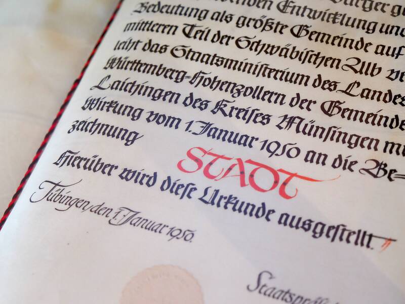 Das Bild zeigt eine historische Urkunde mit kalligraphischem Text in Schwarz und Rot, die auf einer cremefarbenen Unterlage liegt.