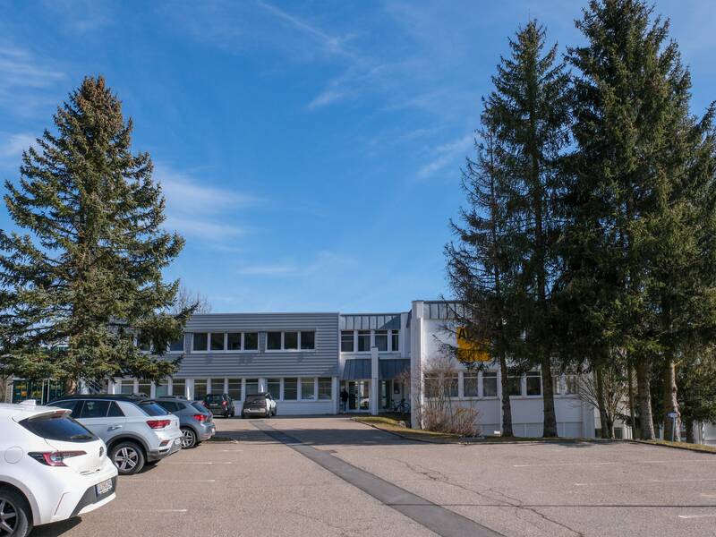 EIn 2 Stöckiges Gebäude umgeben und teilweise verdeckt von Bäumen mit großer geteerter Fläche vor dem mittig liegendem Eingang