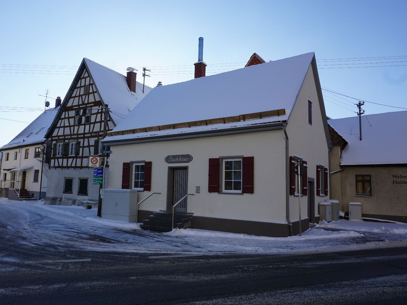 Ein Fachwerkhaus und ein kleines Gebäude mit einem Schild "Backhaus" stehen bei winterlichen Bedingungen an einer verschneiten Straße.