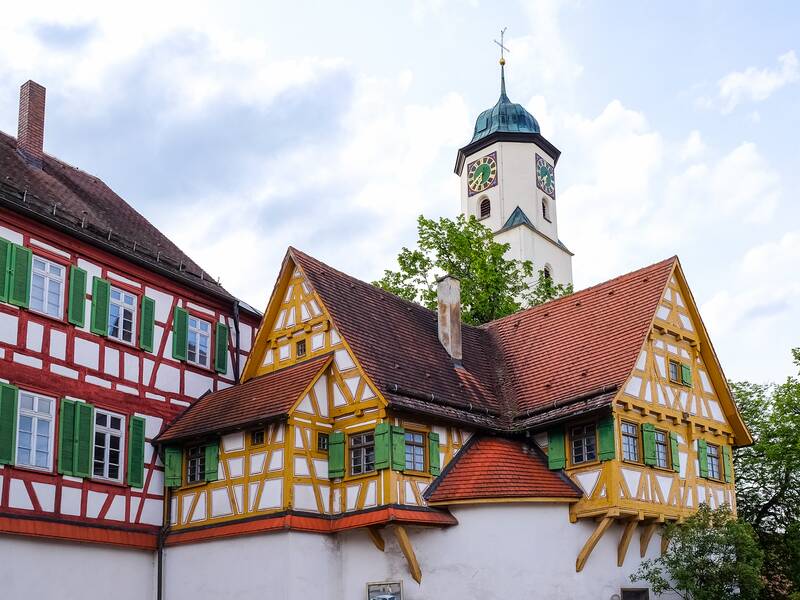 Das Bild zeigt ein historisches Fachwerkhaus mit bunten Balken und grünen Fensterläden vor einem Kirchturm mit einer Uhr, umgeben von Bäumen.