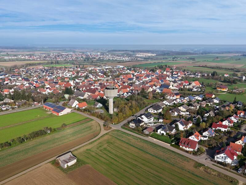 Die Luftaufnahme zeigt eine weitläufige, grüne Landschaft mit einer anscheinend ländlichen Siedlung aus überwiegend roten und weißen Dächern, umgeben von Feldern.