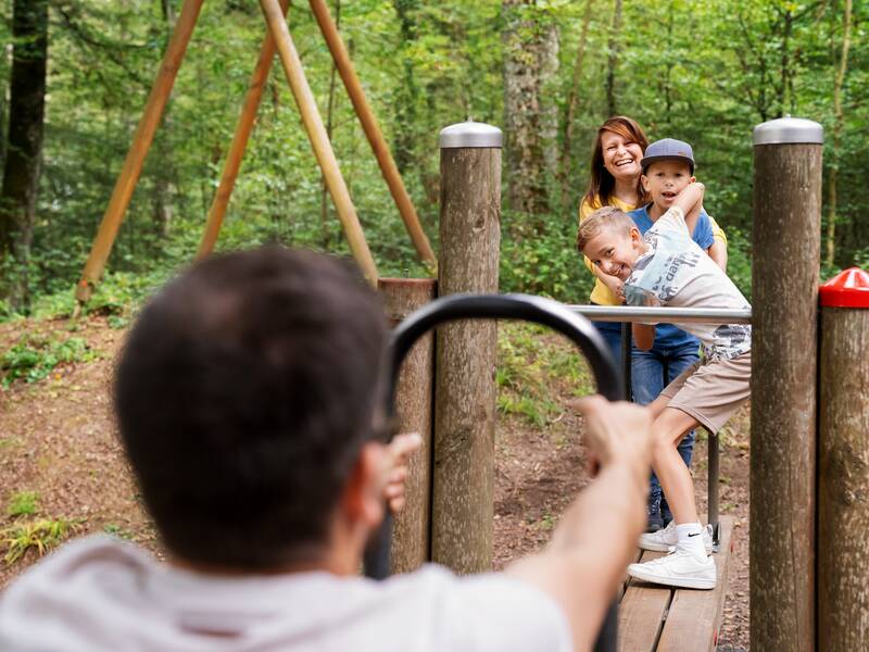 Eine Gruppe von Menschen hat Spaß auf einem Spielplatz im Wald, während eine Person im Vordergrund eine Seilbahn vorwärts zieht.
