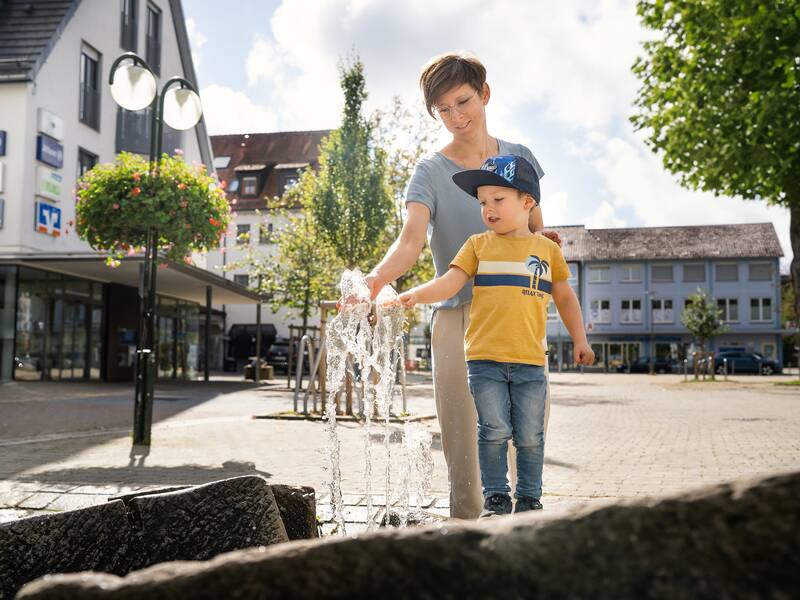 Eine Frau und ein Kind spielen am Leinenweberbrunnen auf einem sonnigen Platz in einer Stadt.