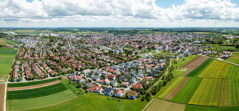 Ein Luftbild zeigt eine weitläufige, grüne Landschaft mit einer Stadt, deren Dächer meist rot und grau sind, umgeben von Feldern und Wiesen unter einem bewölkten Himmel.