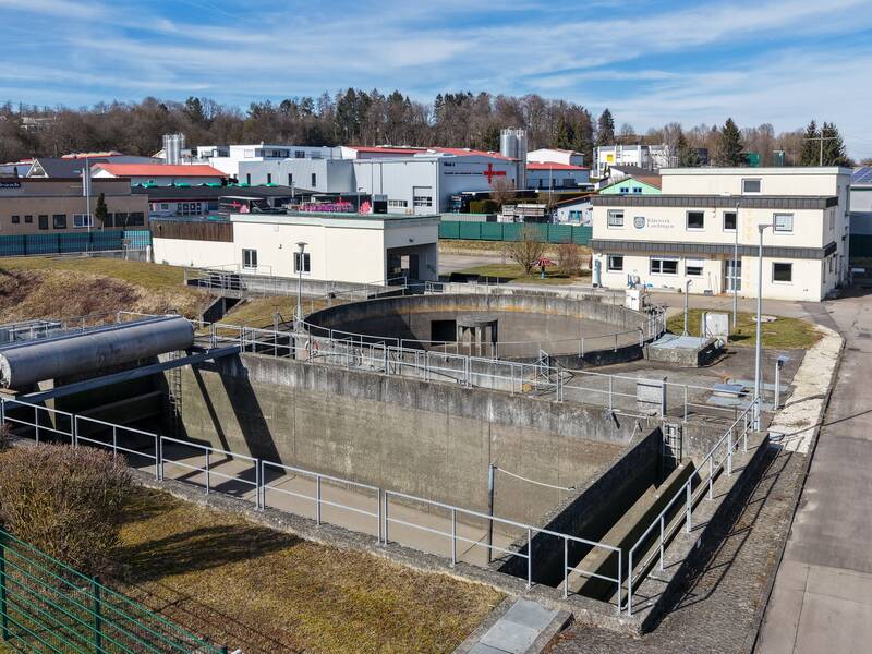 Das Bild zeigt eine Kläranlage mit mehreren Gebäuden, einem runden Becken und umgebendem Gelände unter klarem Himmel.