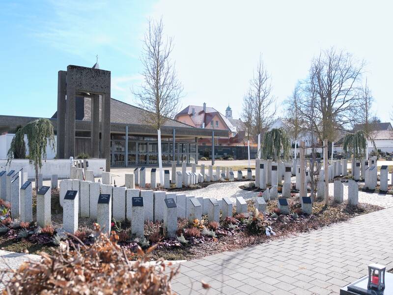 Ein moderner Friedhof mit Gedenksteinen in einem Kreis angeordnet, umgeben von Bäumen und einem Gebäude im Hintergrund.
