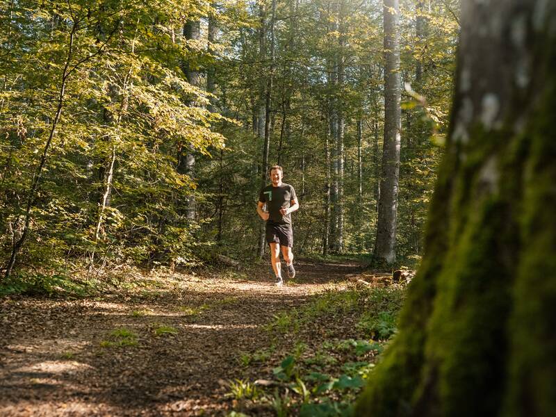 Ein Mann joggt auf einem Waldweg, umgeben von hohen Bäumen und Sonnenlicht, das durch das Blätterdach fällt.