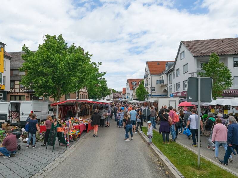 Eine belebte Straße mit zahlreichen Marktständen und Menschen, die Produkte verkaufen und einkaufen, gesäumt von mehrstöckigen Häusern und Bäumen.