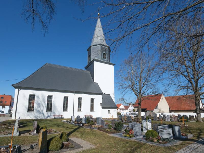 Eine weiße Kirche mit einem grauen Dach steht vor einem klaren blauen Himmel, umgeben von Bäumen und einem Friedhof.