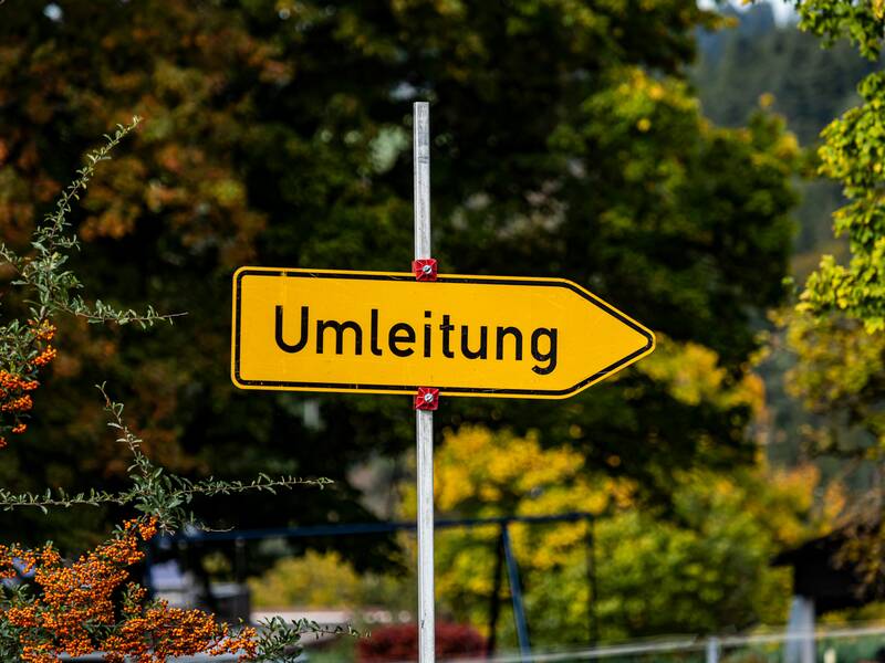 Ein gelbes Umleitungsschild, das in die rechte Richtung zeigt. Im Hintergrund sind herbstliche Bäume uns Sträucher.