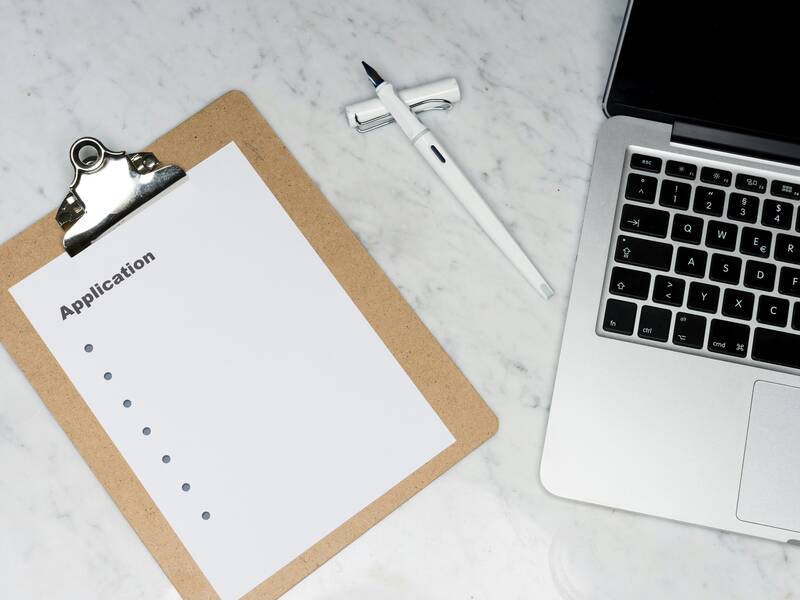 Ein Klemmbrett mit einem leeren, "Application" betitelten Formular liegt neben einem Laptop und einem weißen Stift auf einem hellen Marmortisch.