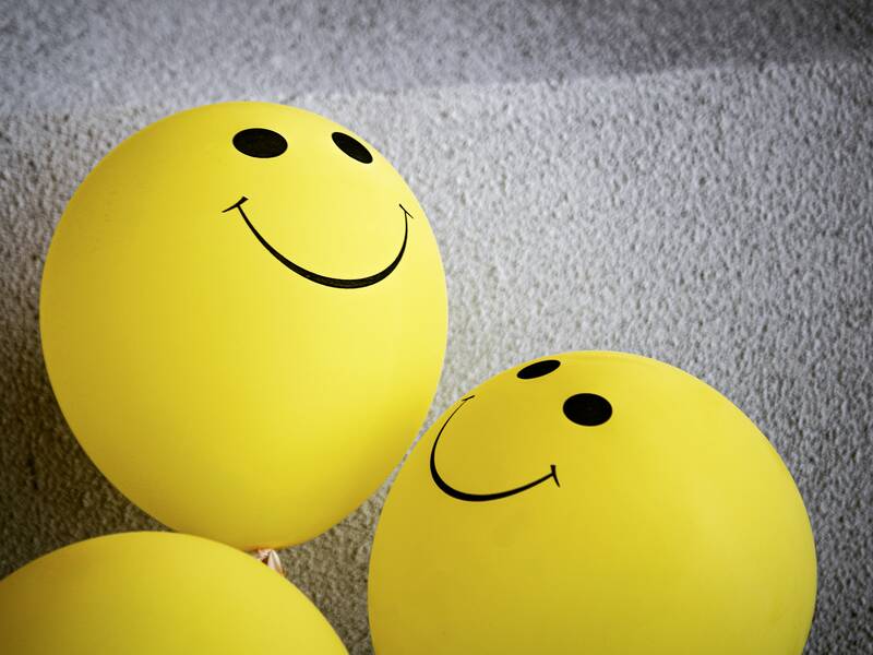 Zwei gelbe Luftballons mit aufgemalten lächelnden Gesichtern schweben vor einem grauen Hintergrund.